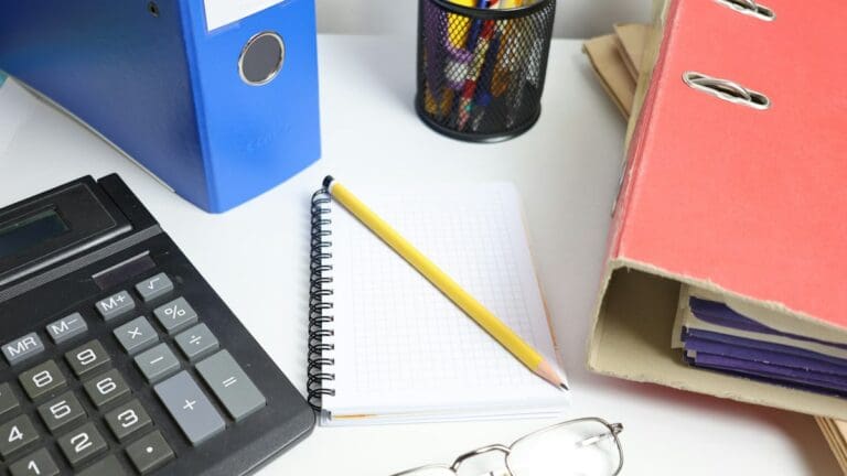 Accountancy desk with blue and red lever-arch files, a desk calculator, a squared notebook with a pencil and reading glasses