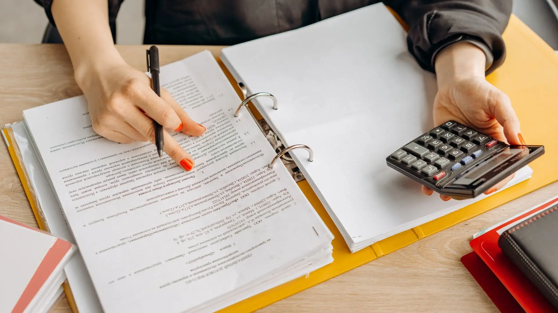 Accountant working through a ring binder of documents, pen in one hand and a desk calculator in the other