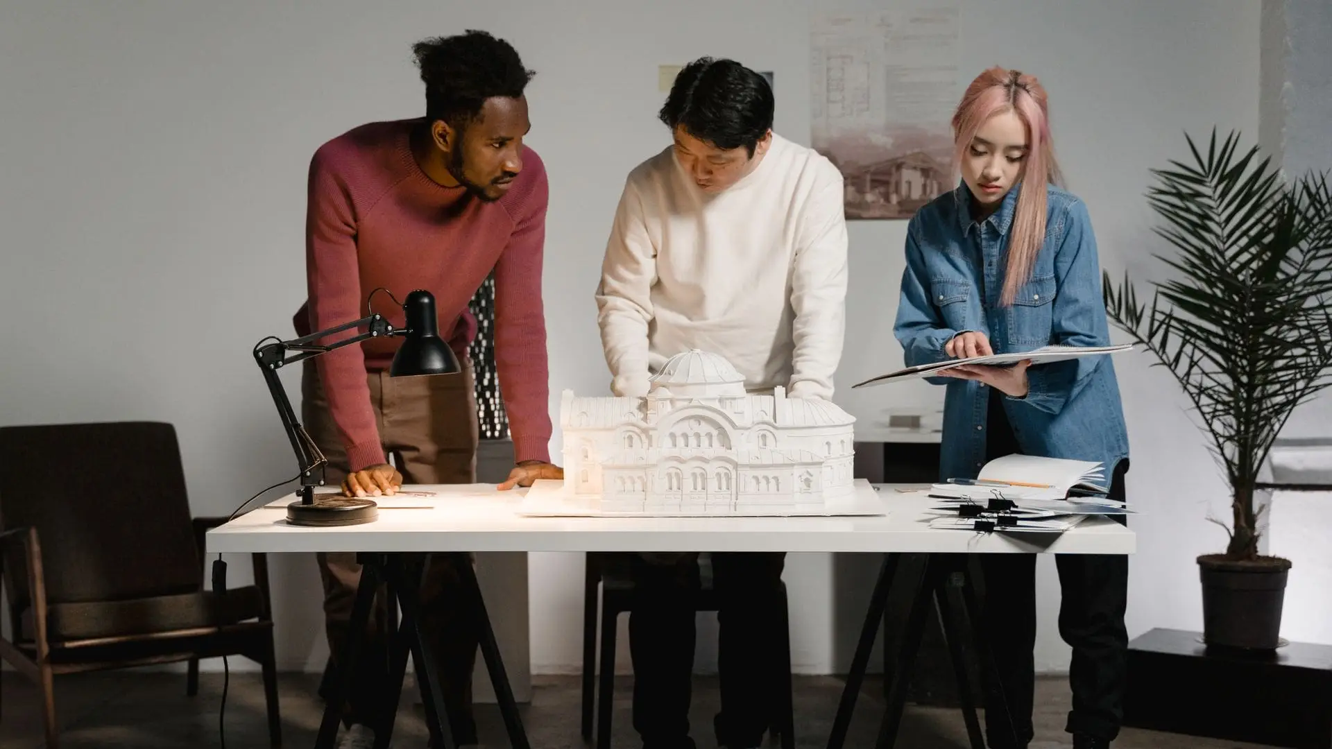 Three architects gathered around a white scale model of a building on a studio table, one holding a set of drawings