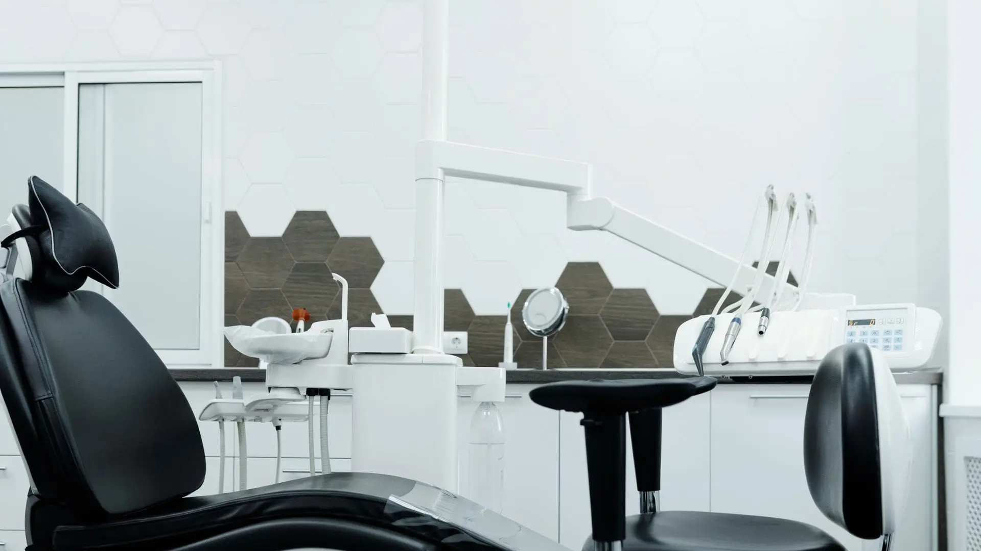 Empty dental surgery with a black patient chair, overhead light arm, instrument tray and a clinician's stool against white and grey hexagonal tiles