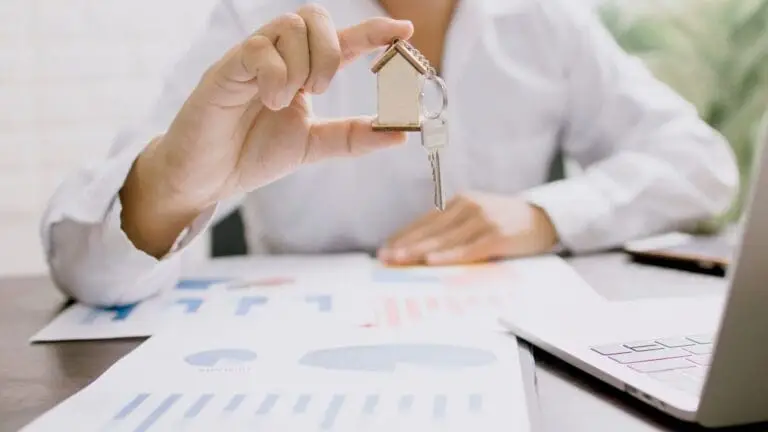 Financial adviser holding a house-shaped key fob over printed mortgage and finance charts beside a laptop