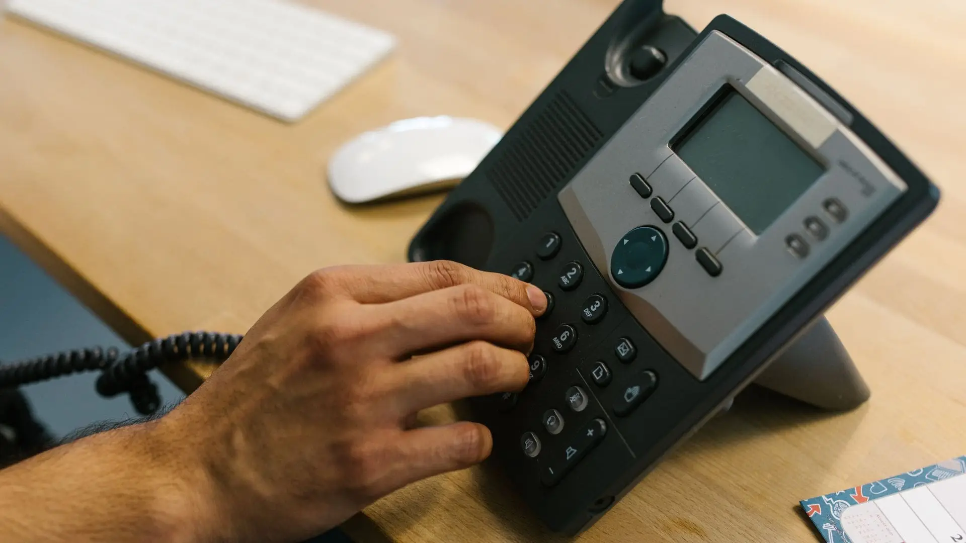 Hand dialling on an IP desk phone on an office desk beside a keyboard and mouse