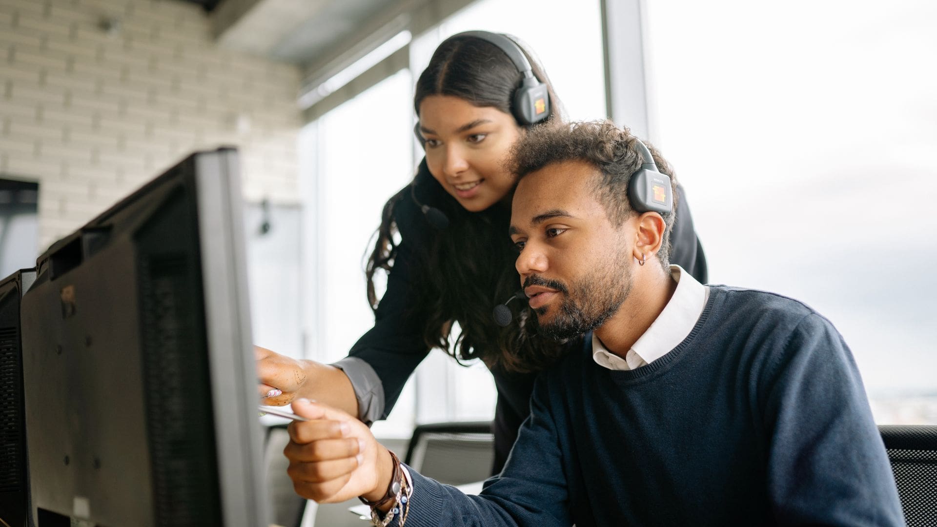 Two helpdesk colleagues wearing headsets working through an issue together at a computer in a bright office