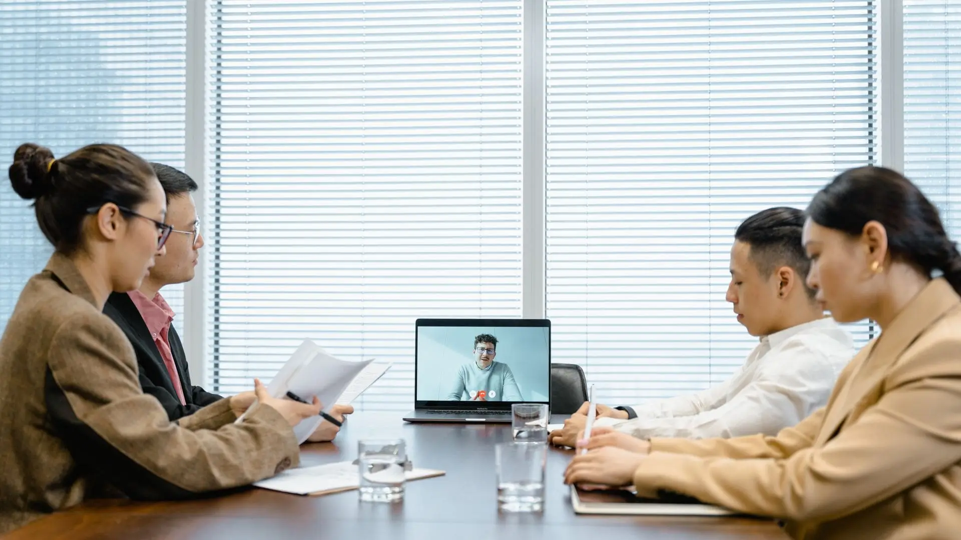 Four colleagues at a meeting room table reviewing documents while a remote colleague joins by video call on a laptop