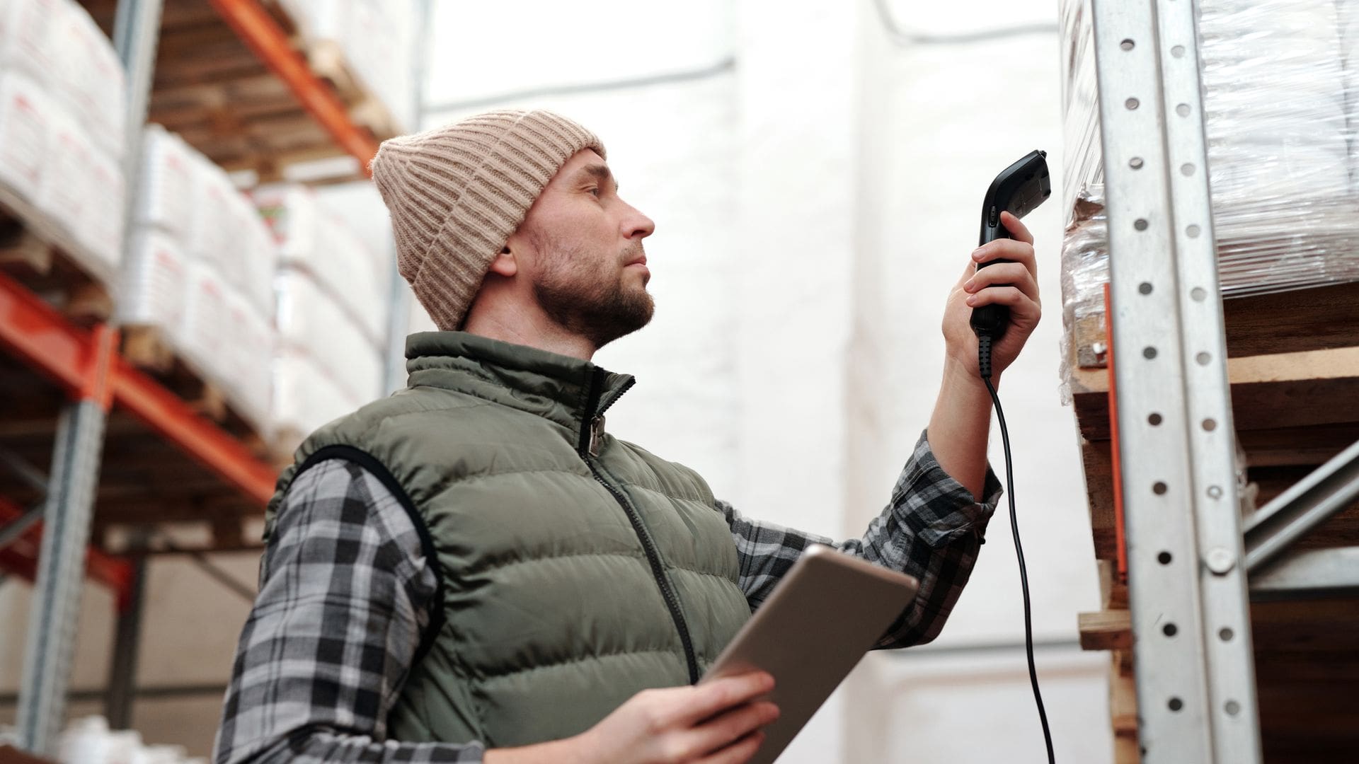 Warehouse worker in a beanie and padded gilet scanning stock on pallet racking with a handheld barcode scanner, holding a tablet