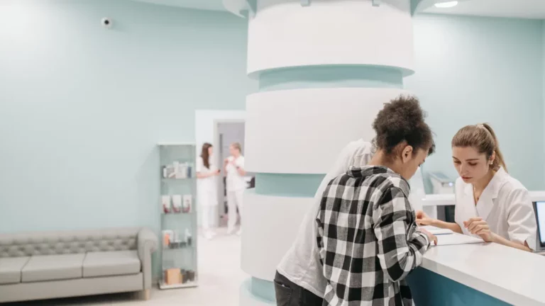Patients checking in with a receptionist at the front desk of a modern medical clinic, with a waiting-area sofa behind them