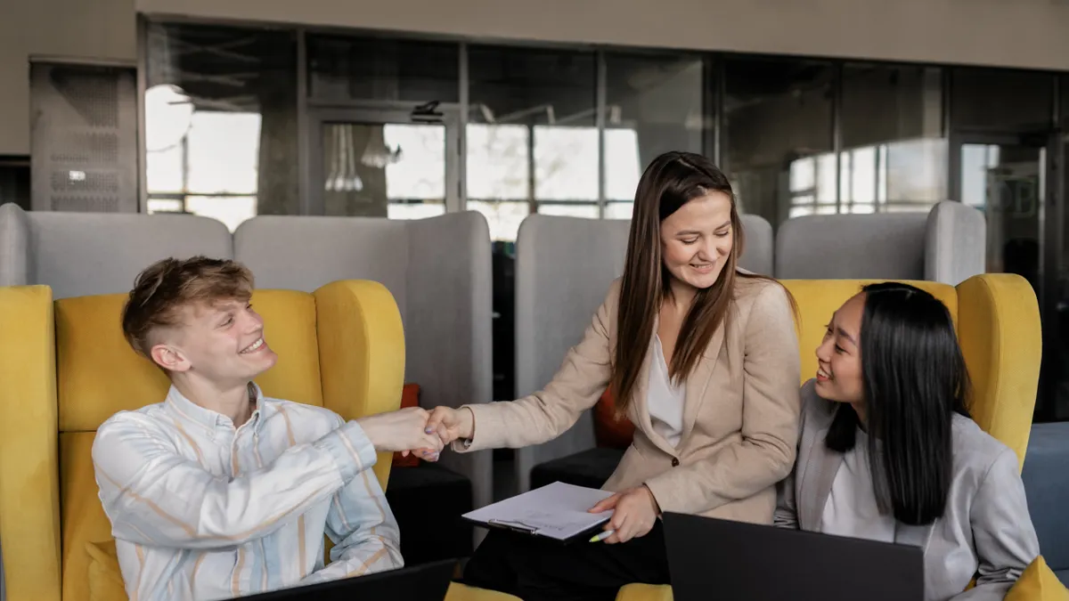 Smiling woman shaking hands with a young man in a bright, modern office while a colleague looks on, welcoming a new client