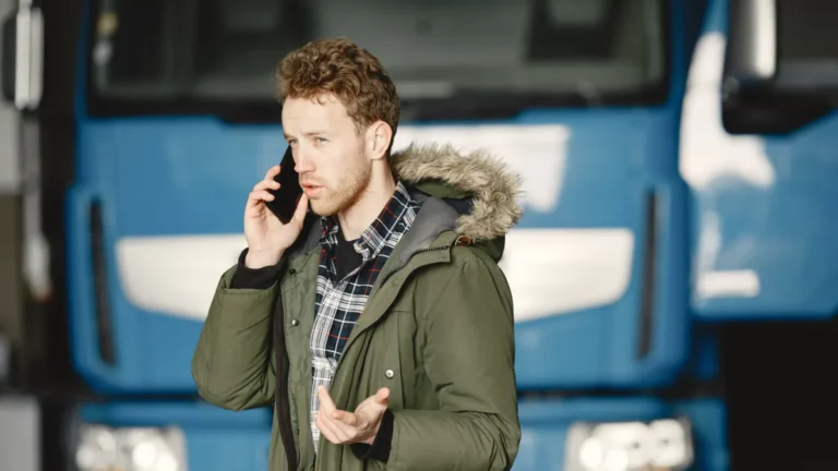Transport manager in a green parka taking a phone call in front of a blue European lorry at a haulage depot